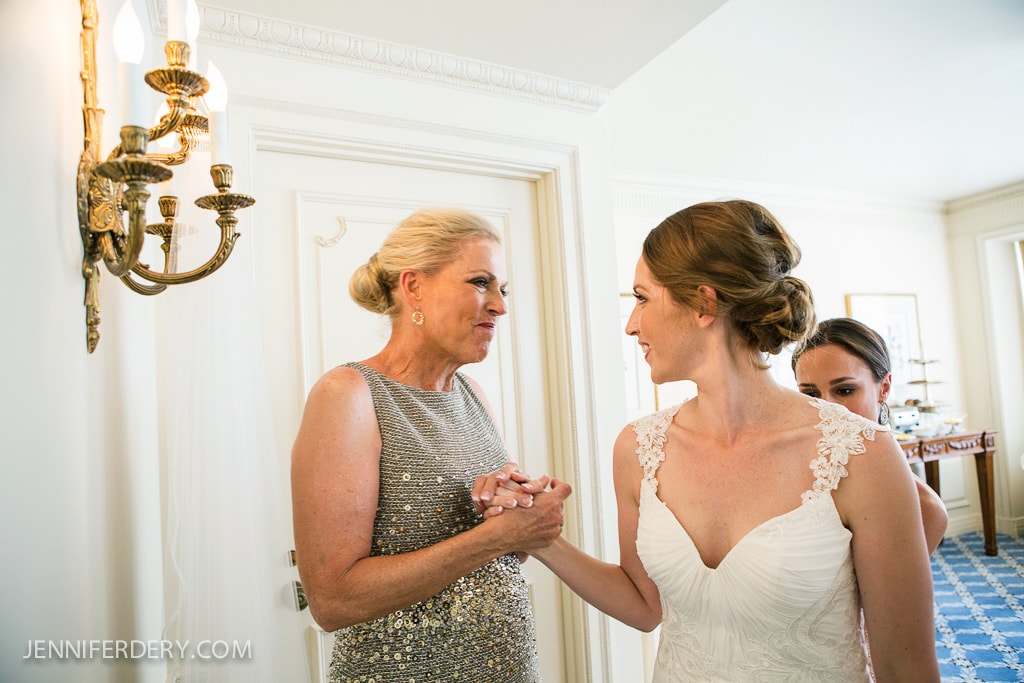A bride in a white lace dress holds hands and smiles with a woman in a sparkly dress. They stand in a well-lit room with elegant decor. Another person is partially visible in the background.
