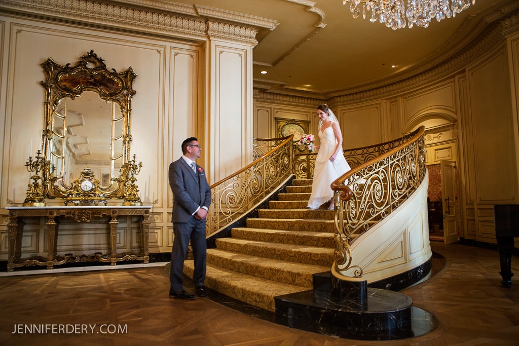 A bride in a white gown and veil descends an ornate curved staircase, holding a bouquet. A groom in a gray suit waits at the bottom. The elegant room features a large mirror, decorative sconces, and a chandelier.