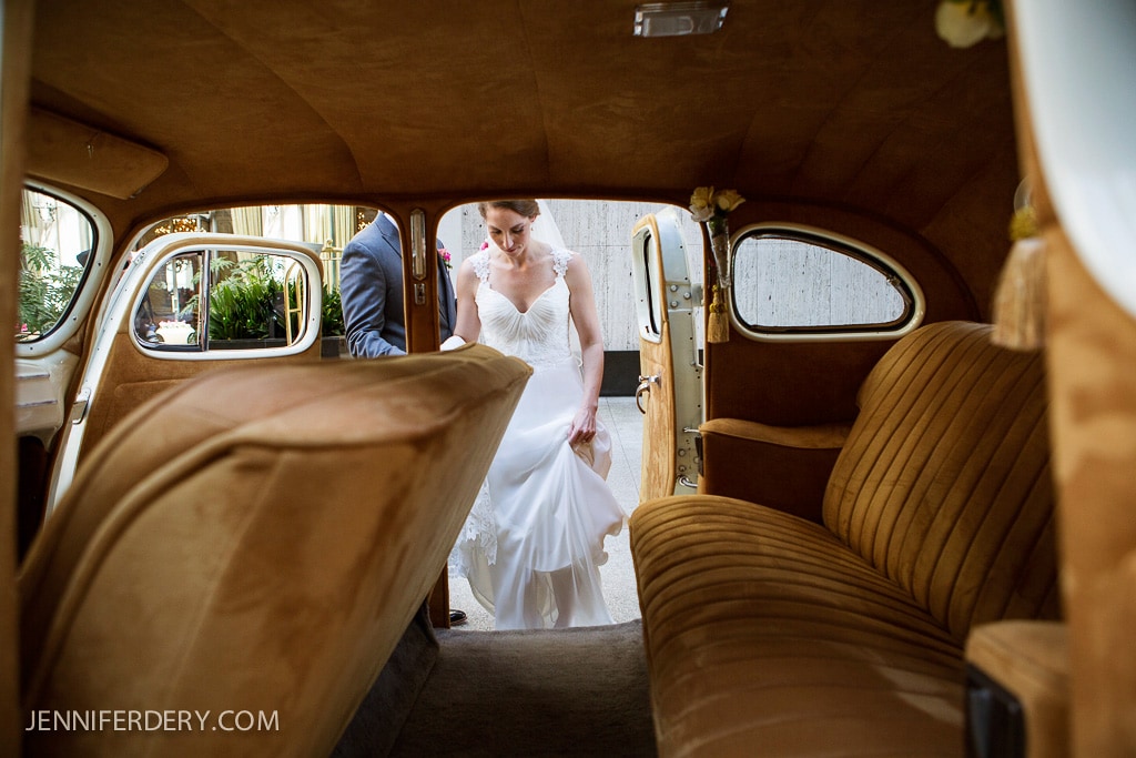 A bride in a white dress prepares to step into a vintage car with a plush, beige interior. The car door is open, and another person is partially visible outside, assisting her. The atmosphere is elegant and classic.