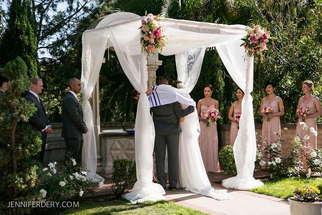 A couple stands under a floral, white canopy during an outdoor wedding ceremony. A person with a shawl embraces them. Bridesmaids in pink dresses and a groomsman in a gray suit stand nearby. Lush greenery surrounds the scene.