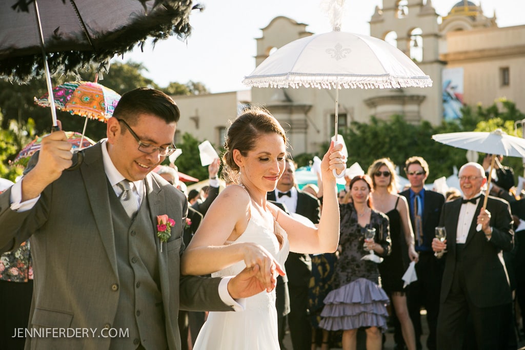 A bride and groom joyfully hold hands, each carrying white parasols. They are surrounded by smiling guests, some holding colorful umbrellas, during an outdoor celebration. A historic building is visible in the background.