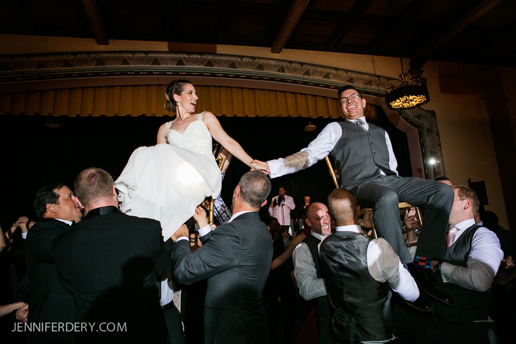 A bride and groom are lifted on chairs during a lively celebration. The groom is wearing a suit, and the bride is in a white gown. Guests in formal attire surround them, smiling and cheering. The setting has a vintage-style interior.