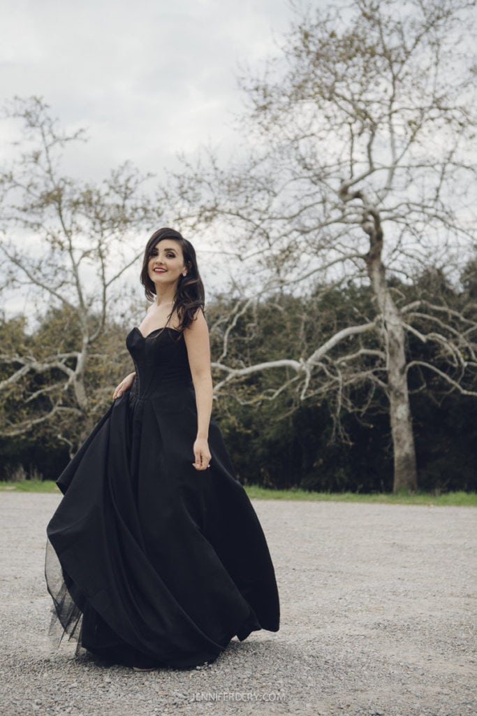 A woman in a flowing black gown stands outdoors on a gravel path. Bare trees and greenery are in the background. The sky is overcast, adding a moody atmosphere to the scene.