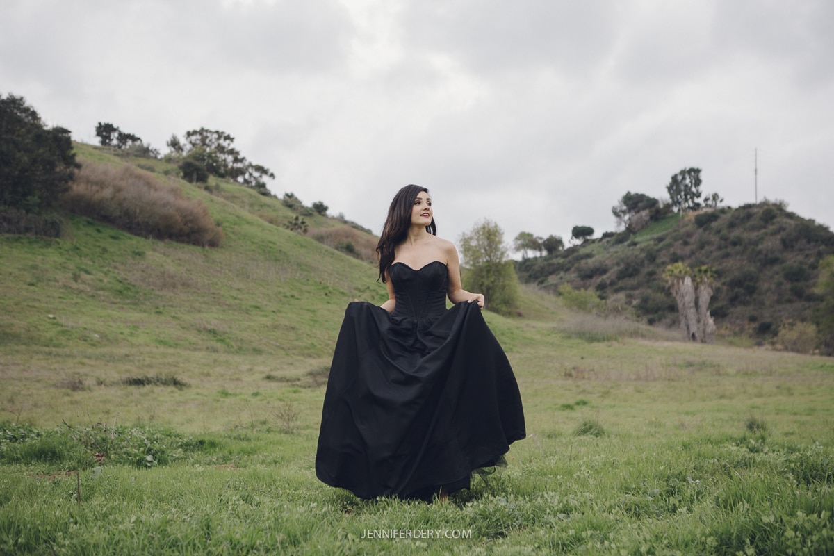 A woman in a flowing black dress stands in a grassy field, holding part of her skirt and gazing to the side. The landscape is green with rolling hills and a cloudy sky.