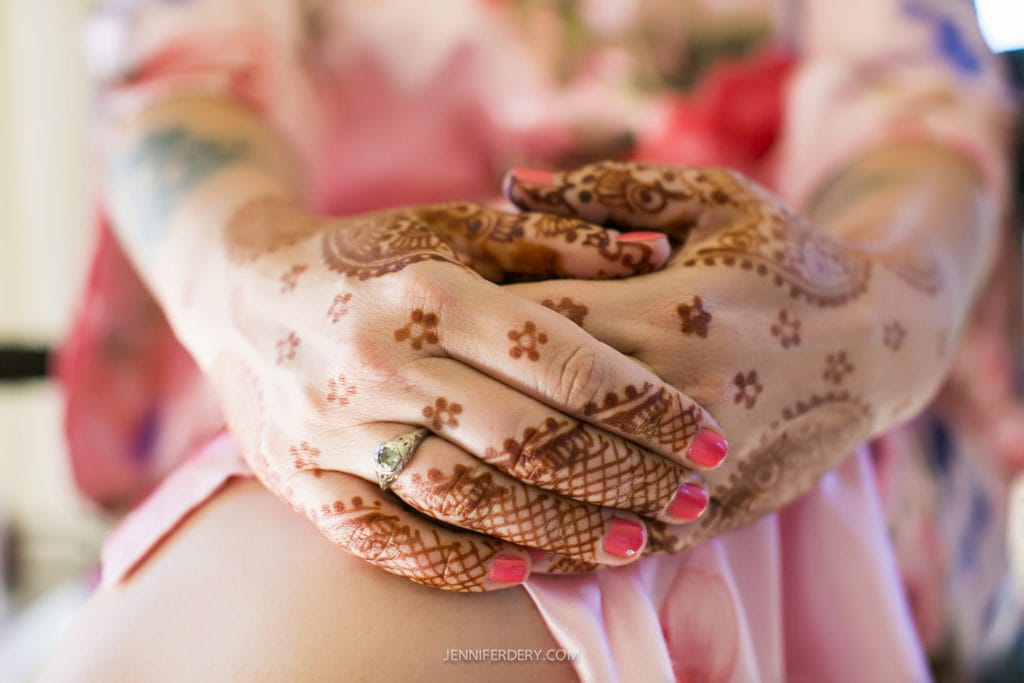 Close-up of folded hands with intricate henna designs and pink nail polish, resting on a pink floral fabric. A silver ring adorns one finger.