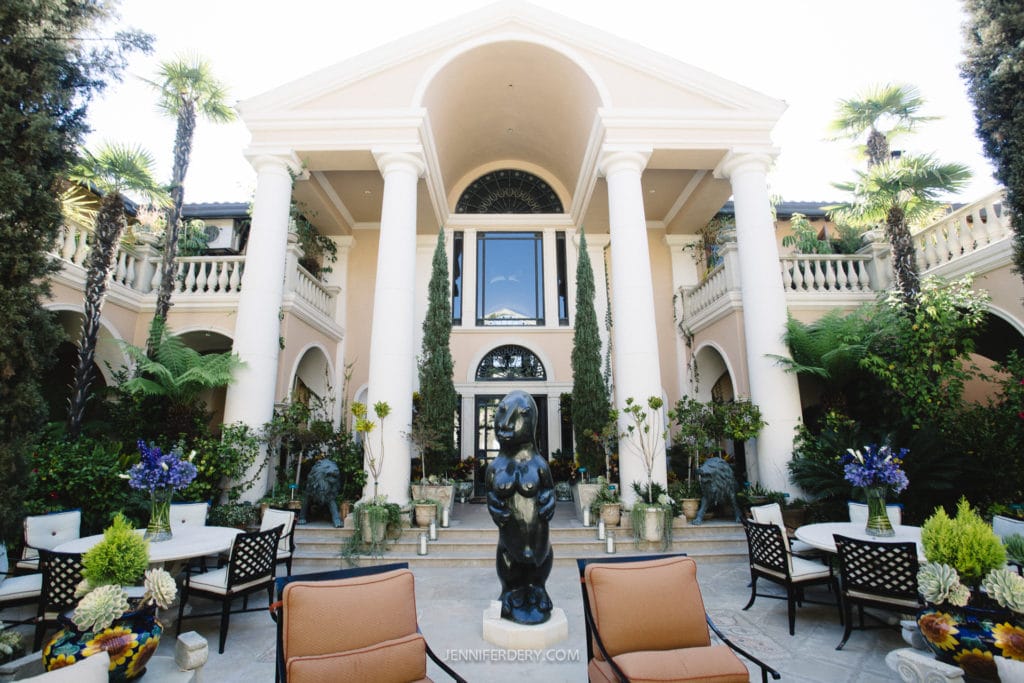 A grand mansion featuring tall white columns and a large central window is surrounded by lush greenery. The patio area includes sculpted statues, flowering plants, and cushioned seating, creating an elegant and inviting outdoor space.