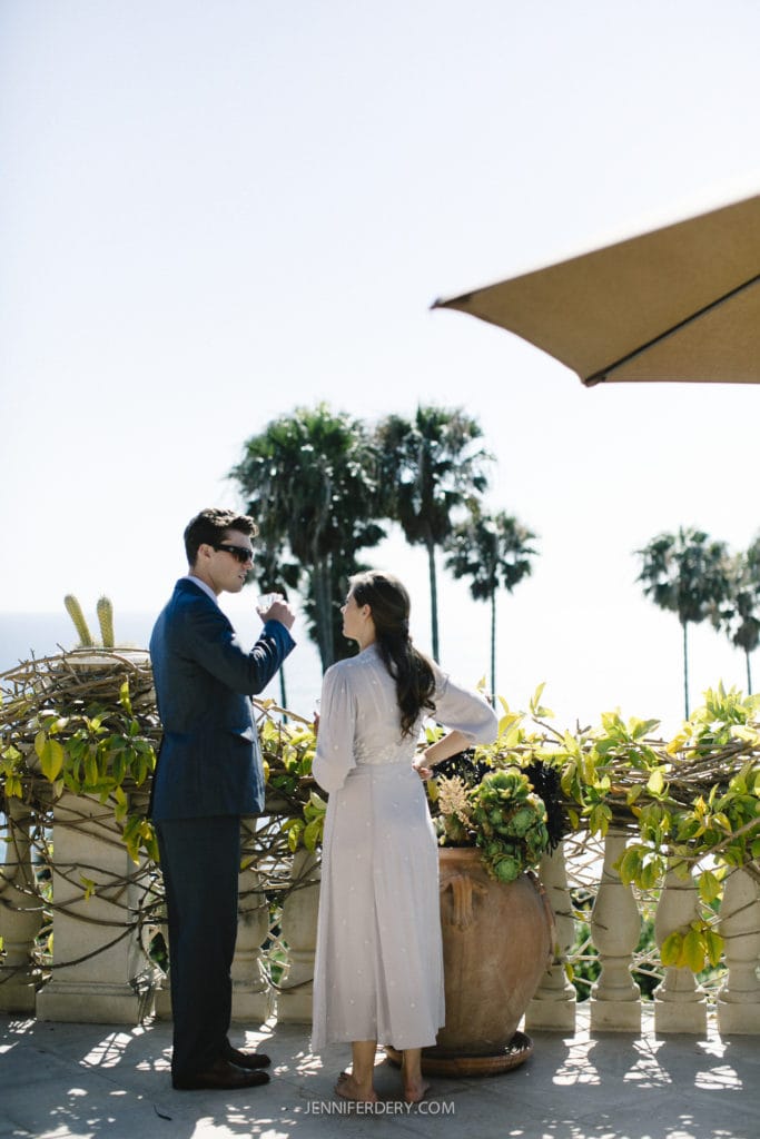 A man in a suit and a woman in a dress stand on a sunlit terrace, engaged in conversation. They overlook an ocean view with palm trees under a clear sky. A large potted plant is nearby, and part of a sunshade is visible above them.