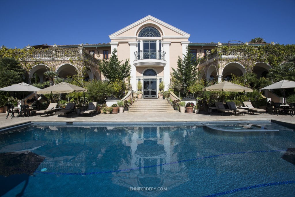 A luxurious villa with a grand entrance and arched balconies is reflected in a large, clear swimming pool. The area is surrounded by lush greenery and sun loungers with umbrellas, under a clear blue sky.