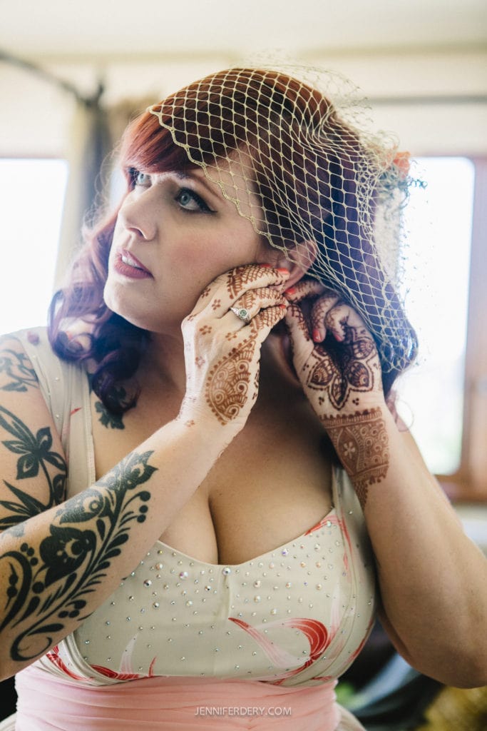 A woman with red hair adjusts her earring. She is wearing a white dress with pink accents and a netted headpiece. Her arms and hands are adorned with intricate henna designs, and she is indoors with natural light coming through the window.