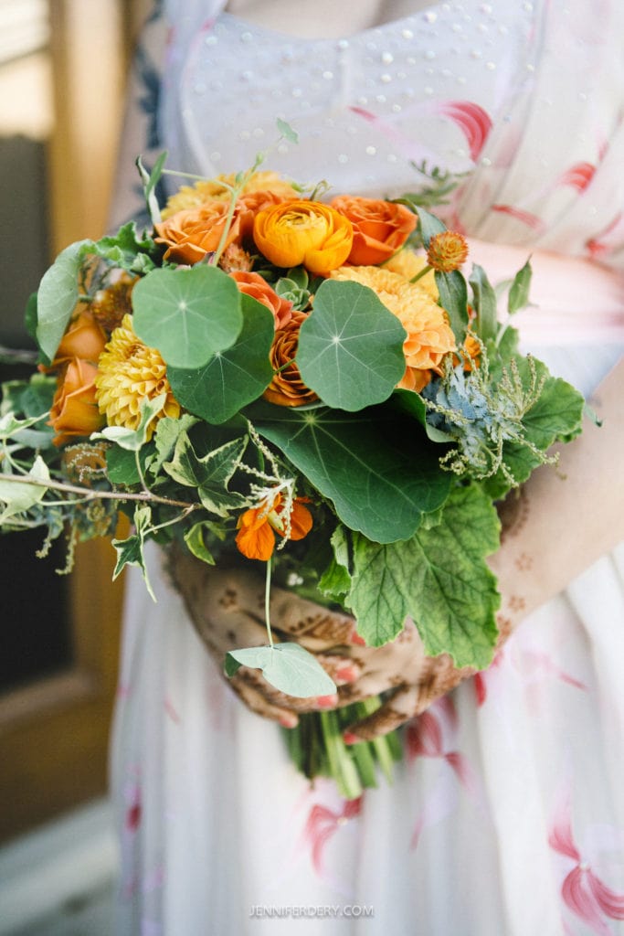 A person holding a vibrant bouquet of orange and yellow flowers, including ranunculus and dahlias, mixed with green leaves. The background features a blurred dress with pink floral patterns and detailed henna on the hands.