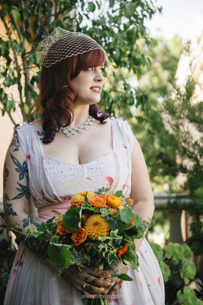 A woman in a white and pink vintage dress and small veil holds a bouquet of orange and yellow flowers. She stands outdoors with a background of greenery and trees, looking to the side.