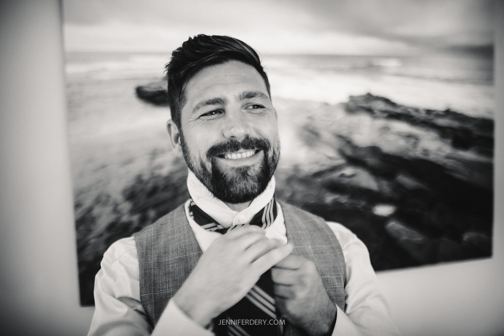 A man with a beard smiles while adjusting his tie. He is wearing a vest over a collared shirt. A blurred ocean landscape is visible in the background.