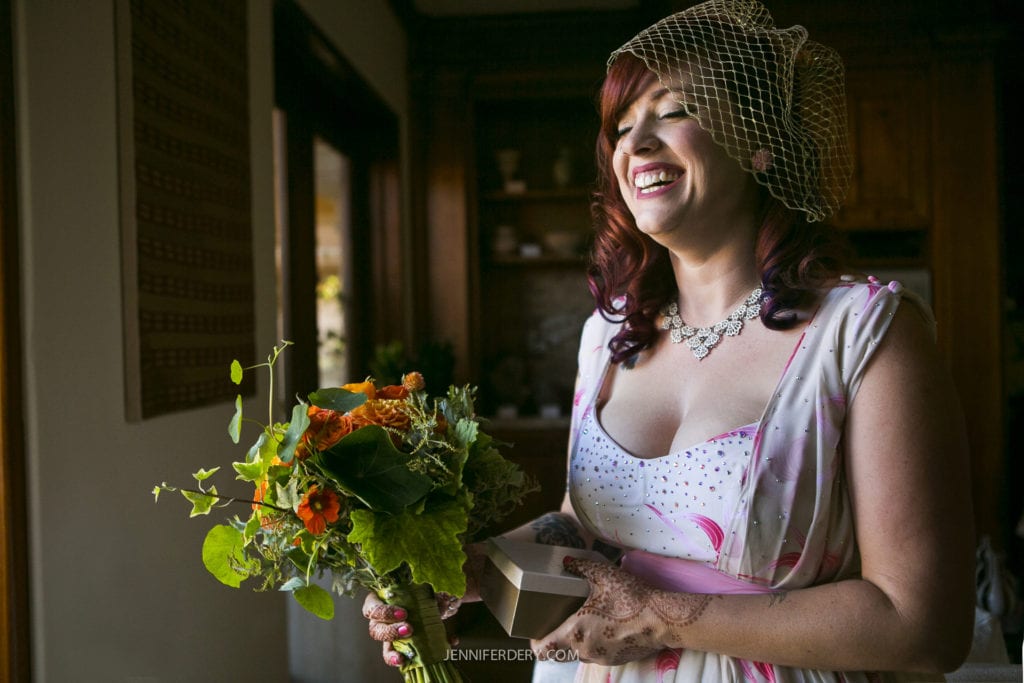 A joyful bride with red hair, wearing a netted veil and holding a vibrant bouquet, stands indoors. She is dressed in a white dress with a pink ribbon and floral patterns, and she wears an elegant necklace. Sunlight softly illuminates the room.