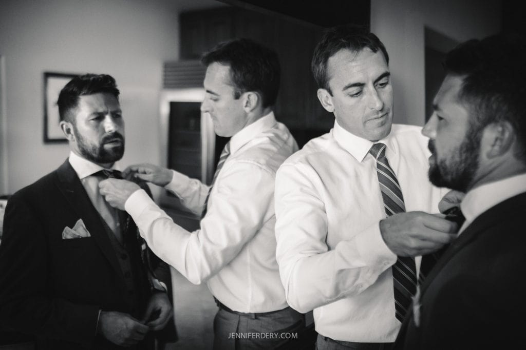 A black and white image of four men in a room. Two men are helping the other two adjust their ties. They are dressed in suits, preparing for a formal event. The scene conveys a sense of camaraderie and attention to detail.