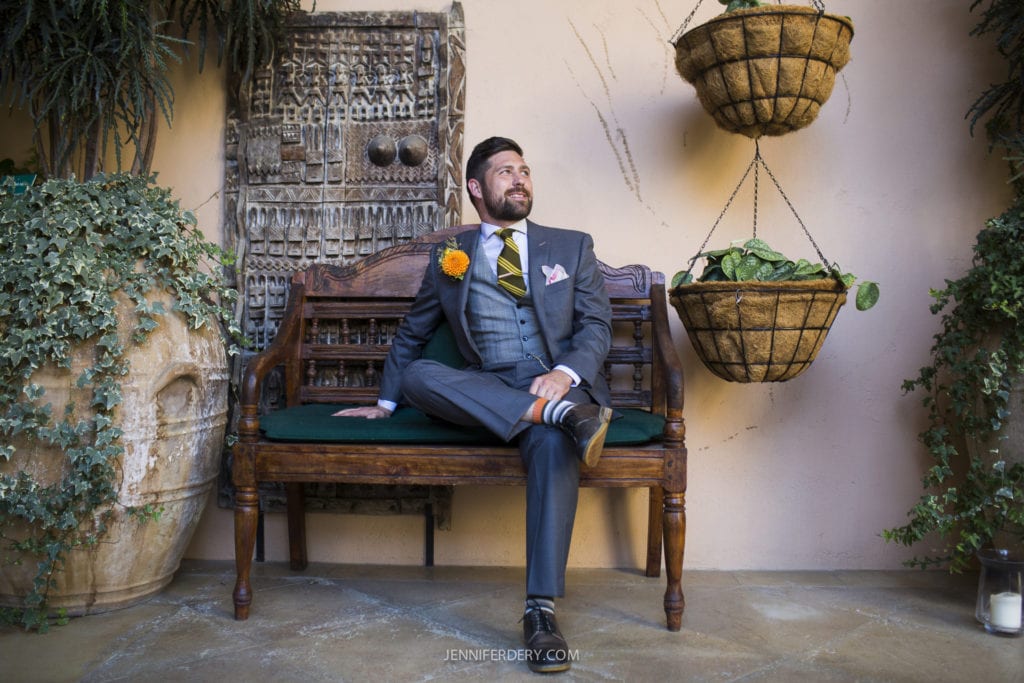 A man in a tailored suit with a yellow boutonniere sits on a wooden bench. He is looking to the side, surrounded by decorative plants and hanging baskets against an ornate wall backdrop.