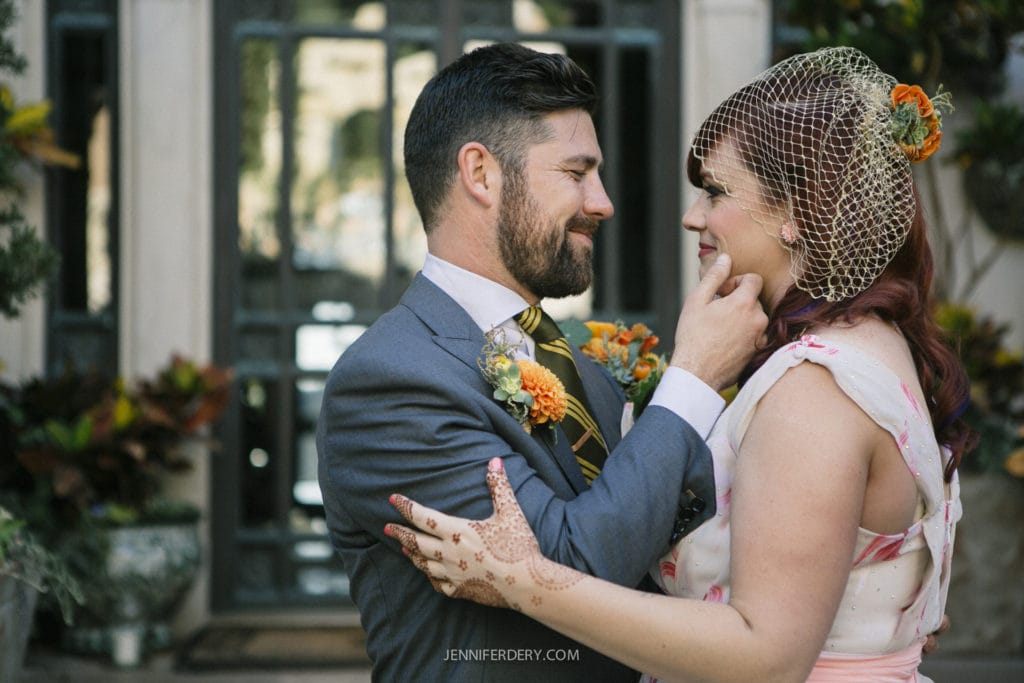 A couple stands close together, smiling at each other. The man is wearing a gray suit and yellow tie with a flower boutonniere, and the woman is in a white dress with a veil and henna on her hand. They are outdoors in front of a building.