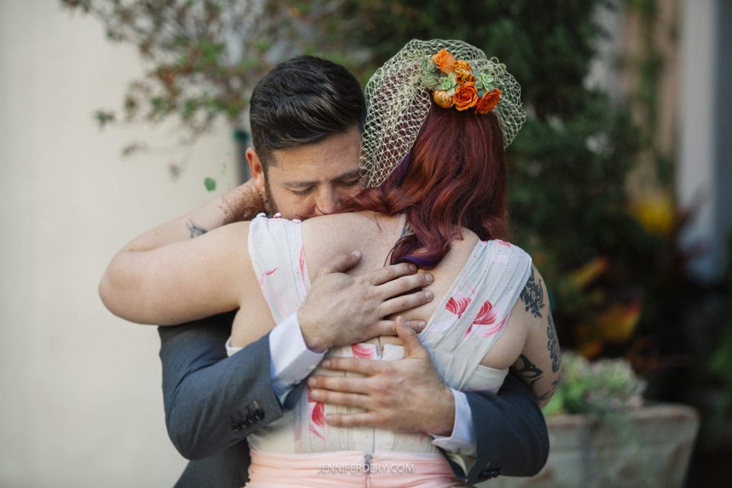 A groom in a gray suit embraces a bride in a floral dress and birdcage veil with orange flowers. They hug tenderly in an outdoor setting, surrounded by plants and soft lighting.