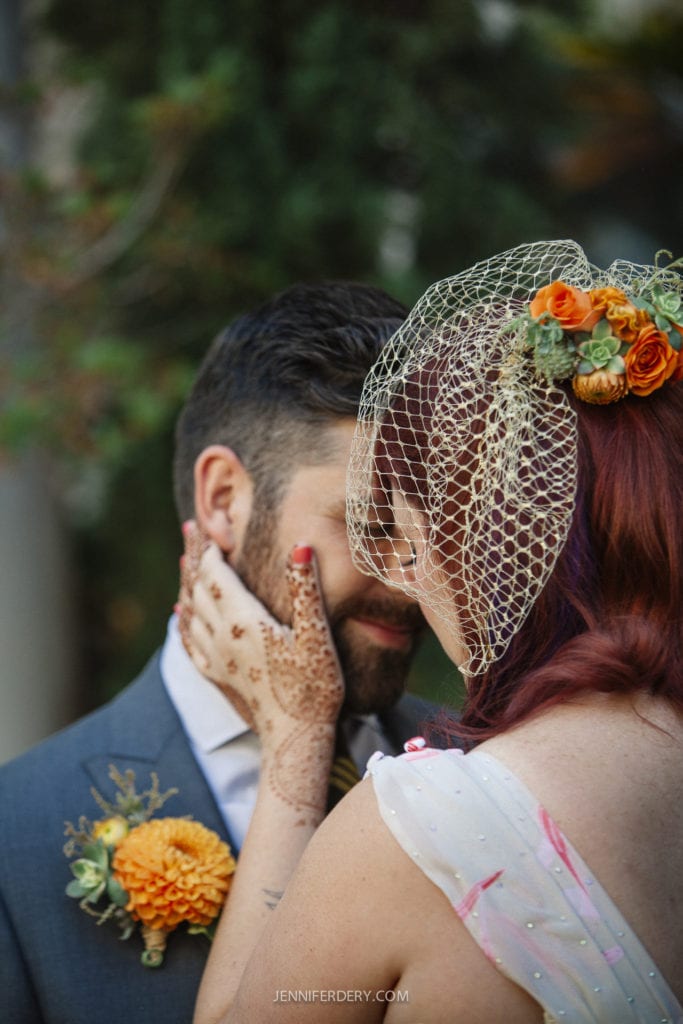 A bride with henna on her hands and a floral hairpiece lovingly touches the face of a groom in a gray suit, who is also wearing a flower on his lapel. A netting veil partially covers her face. Both are standing outdoors.