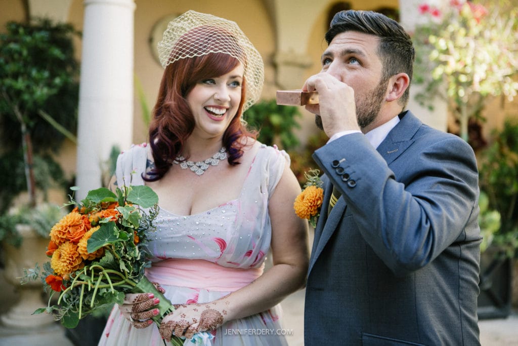 A woman in a vintage-style dress holding a bouquet with orange flowers stands next to a man in a suit, who is blowing into a pink kazoo. They are outdoors, smiling and enjoying the moment in a garden setting.