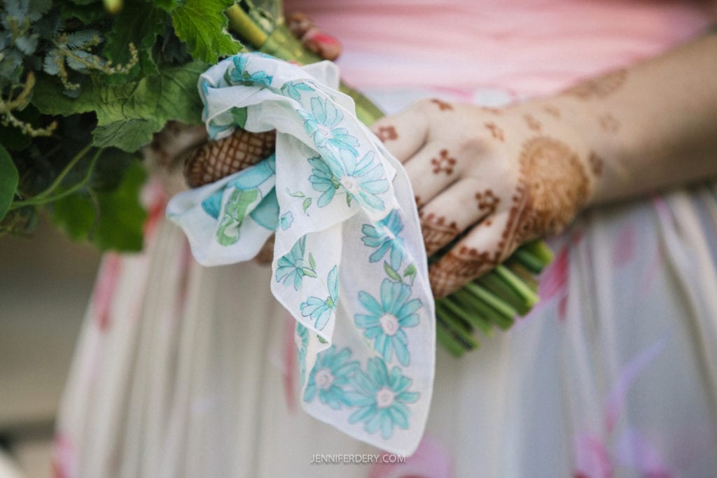 A person holds a bouquet adorned with henna-decorated hands. A blue floral handkerchief is tied around the bouquet. The person wears a pink and white floral outfit.