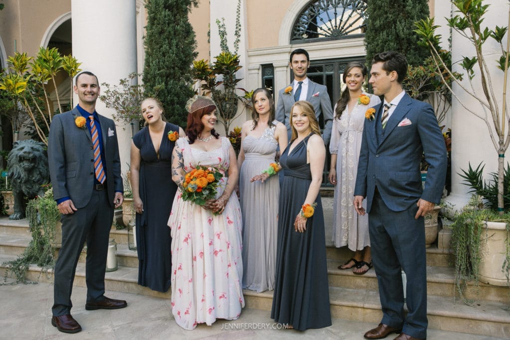 A bride in a floral gown holds a bouquet while surrounded by bridesmaids in gray dresses and groomsmen in gray suits with orange accents. They are making silly faces while standing on steps in front of a building with large columns and greenery.