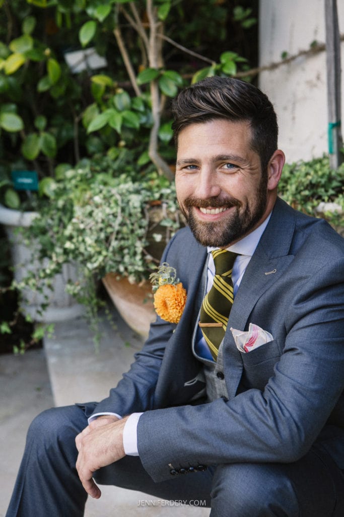 A man in a gray suit and striped tie sits outside, smiling, with greenery in the background. He has a boutonniere with an orange flower and a pocket square.