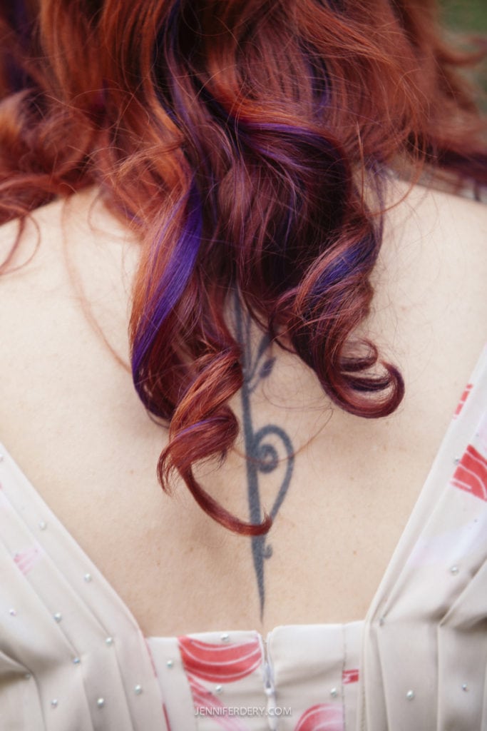A person with long, curly red hair accented with purple highlights, shown from the back. They are wearing a light-colored dress with delicate patterns and have a tattoo with a musical note design on their upper back.