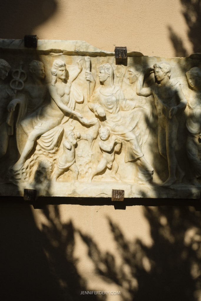 Ancient stone relief depicting several figures, including a seated person receiving a scroll. The artwork is partially in shadow, and the background wall is beige.