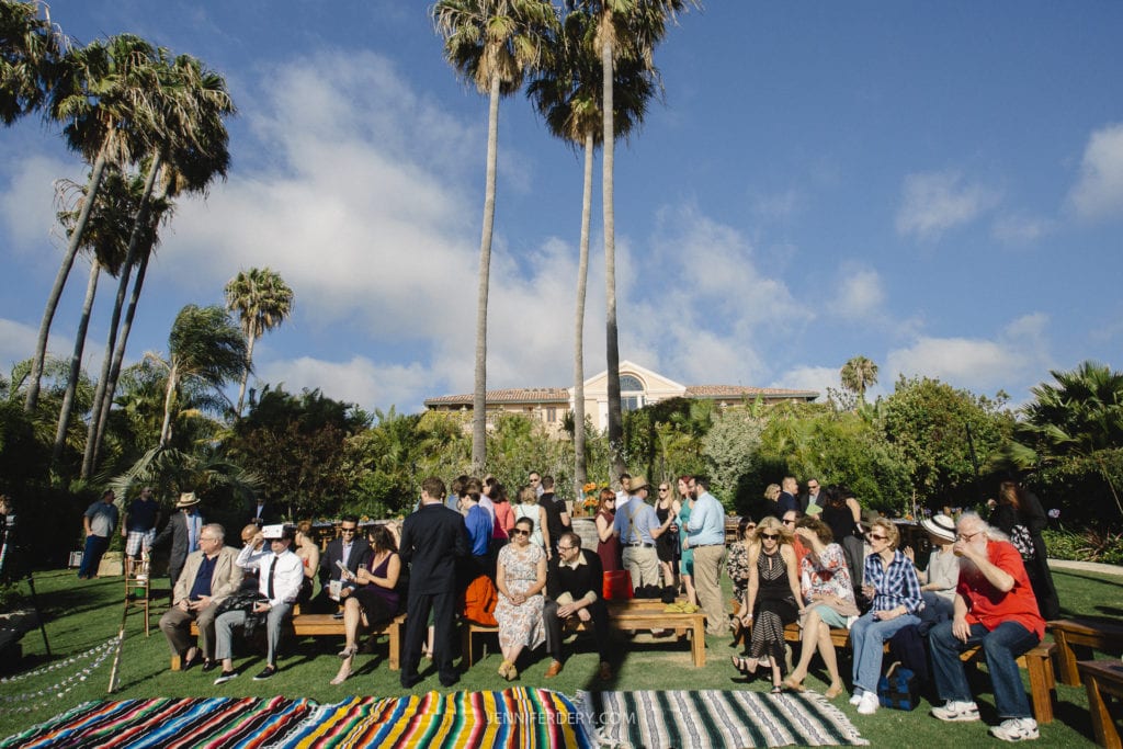 A diverse group of people gathered outdoors, sitting on benches and standing on a lawn. Tall palm trees and a large building in the background. Colorful striped blankets on the ground enhance the festive atmosphere.