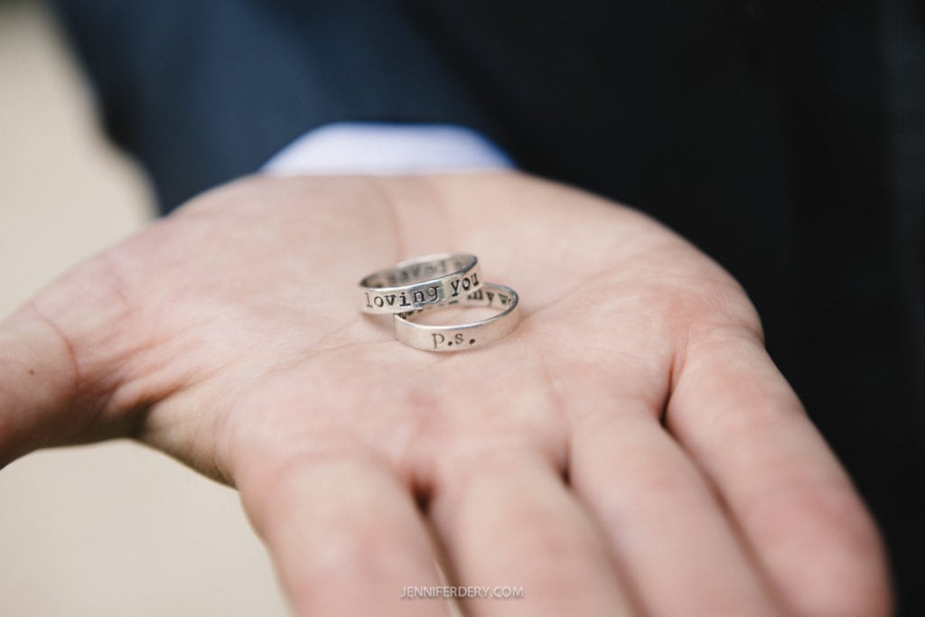 A hand holding two silver rings, one engraved with "loving you always" and the other with "P.S." The background is blurred, highlighting the rings.