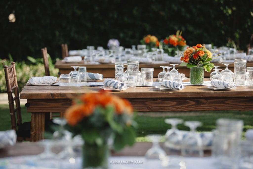 A wooden outdoor table is set for an event, adorned with floral centerpieces, wine glasses, and neatly rolled napkins. The scene is surrounded by greenery, creating a natural and elegant ambiance.