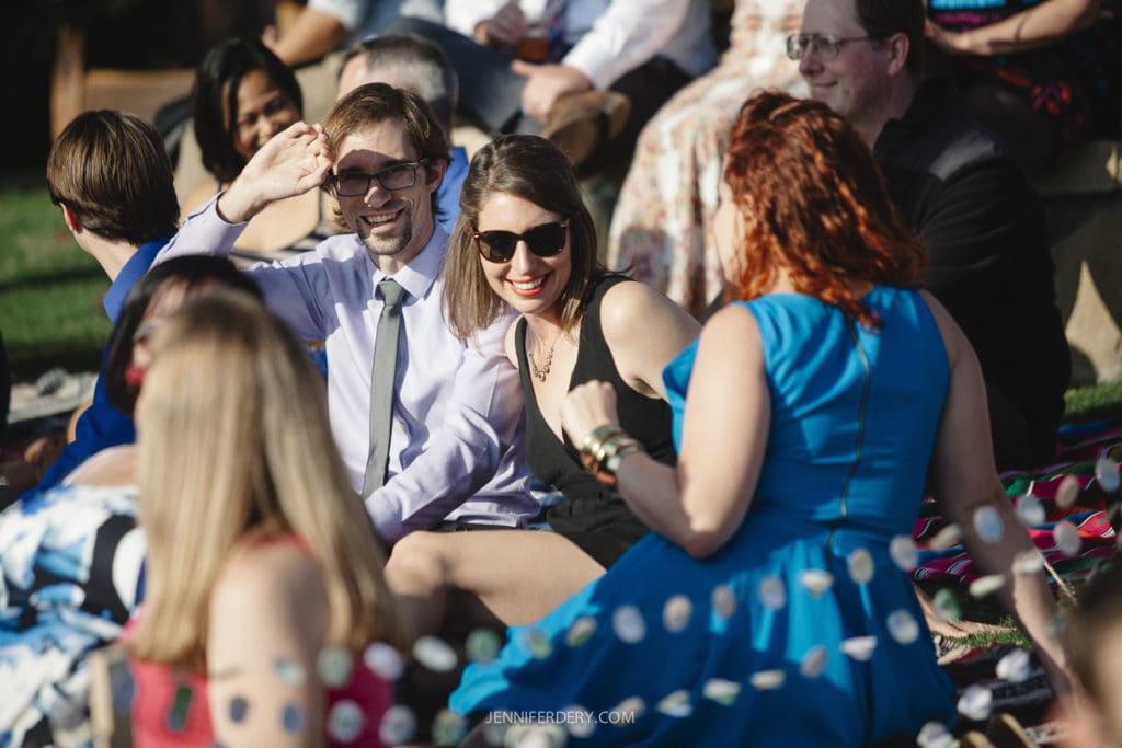 A group of people sitting on colorful blankets outdoors, smiling and chatting. One man in a white shirt and tie is shielding his eyes from the sun, while a woman in sunglasses and a black dress sits beside him. The scene is bright and sunny.