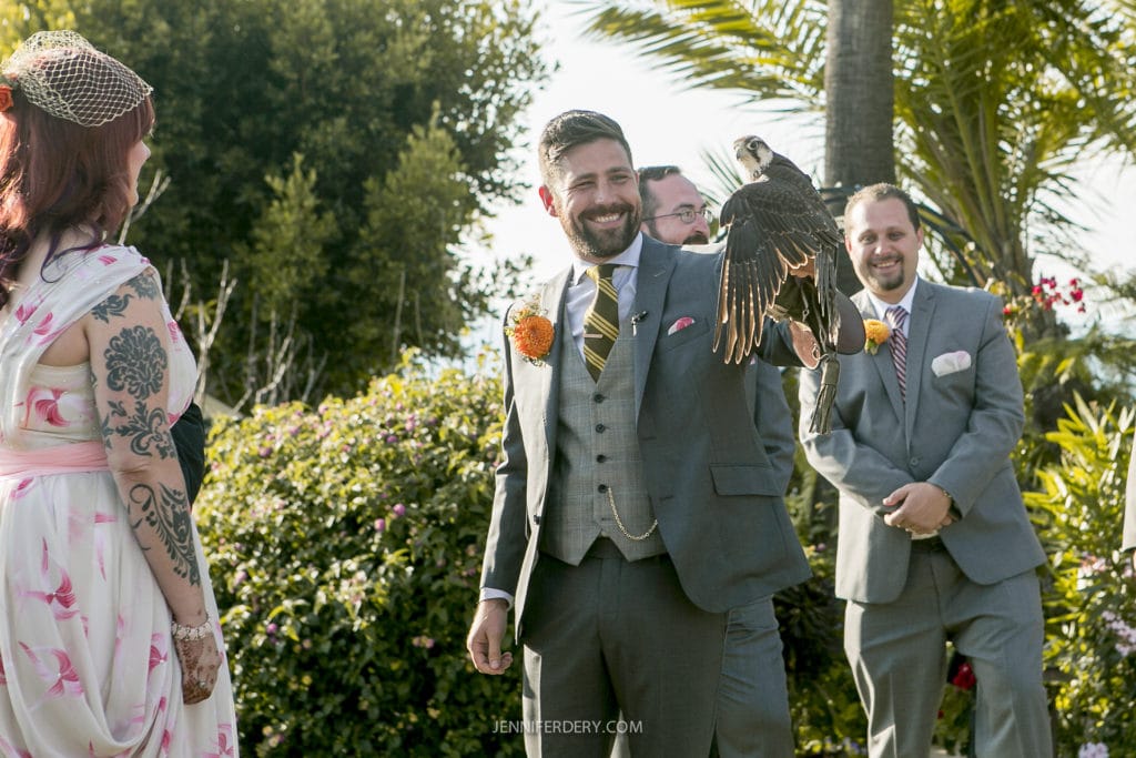 A groom in a grey suit holds a falcon ring bearer on his arm during an outdoor wedding ceremony. The bride, in a flower-patterned dress, watches with guests standing nearby. The setting is sunny, with lush greenery and palm trees in the background.