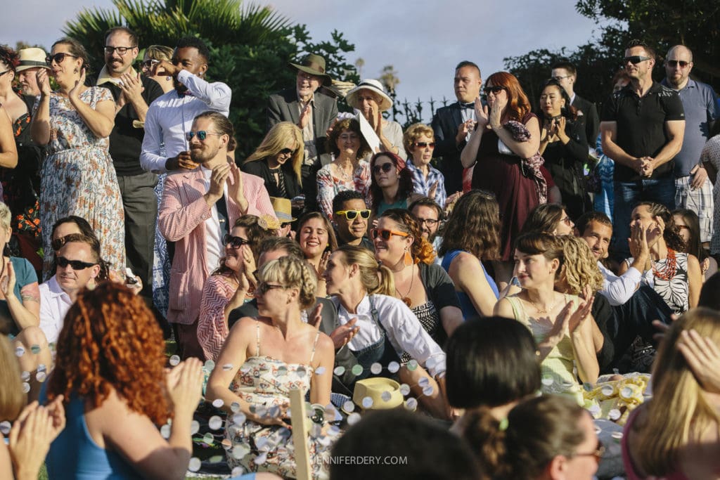 A large group of wedding guests are seated and standing outdoors, wearing a variety of colorful, vintage-style clothing. Some are clapping, while others look on with smiles. Trees and a clear sky are visible in the background.