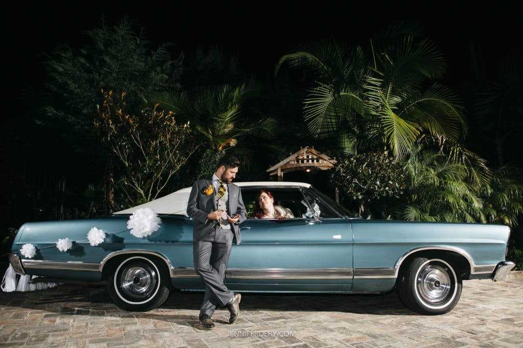 A man in a suit leans against a vintage blue convertible, while a woman sits inside. The car is decorated with white floral accents. They are outside, with lush greenery in the background, and it's nighttime.