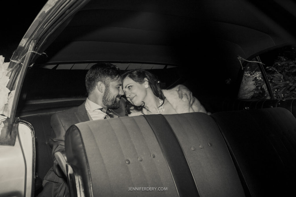 A newlywed couple sits in the backseat of a car, leaning towards each other and smiling. The setting is cozy and intimate, captured in soft black and white. The woman wears a fur shawl, and the man is in a suit.
