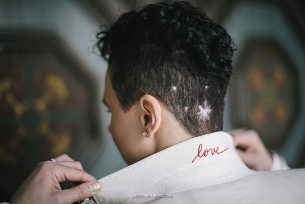 Close-up photo of a person with short curly hair. Visible on their tan collar is the word "love" written in red cursive script. The setting appears to be a casual environment, possibly before or after a Balboa Park wedding.