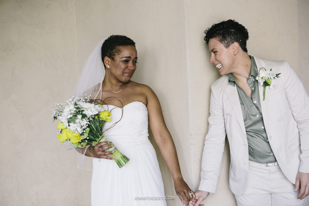 A newlywed couple in wedding attire stands side by side, holding hands and leaning against a wall at Balboa Park. One person is wearing a white dress and holding a bouquet of yellow and white flowers, smiles at her partner. The partner, in a light-colored suit, smiles back.