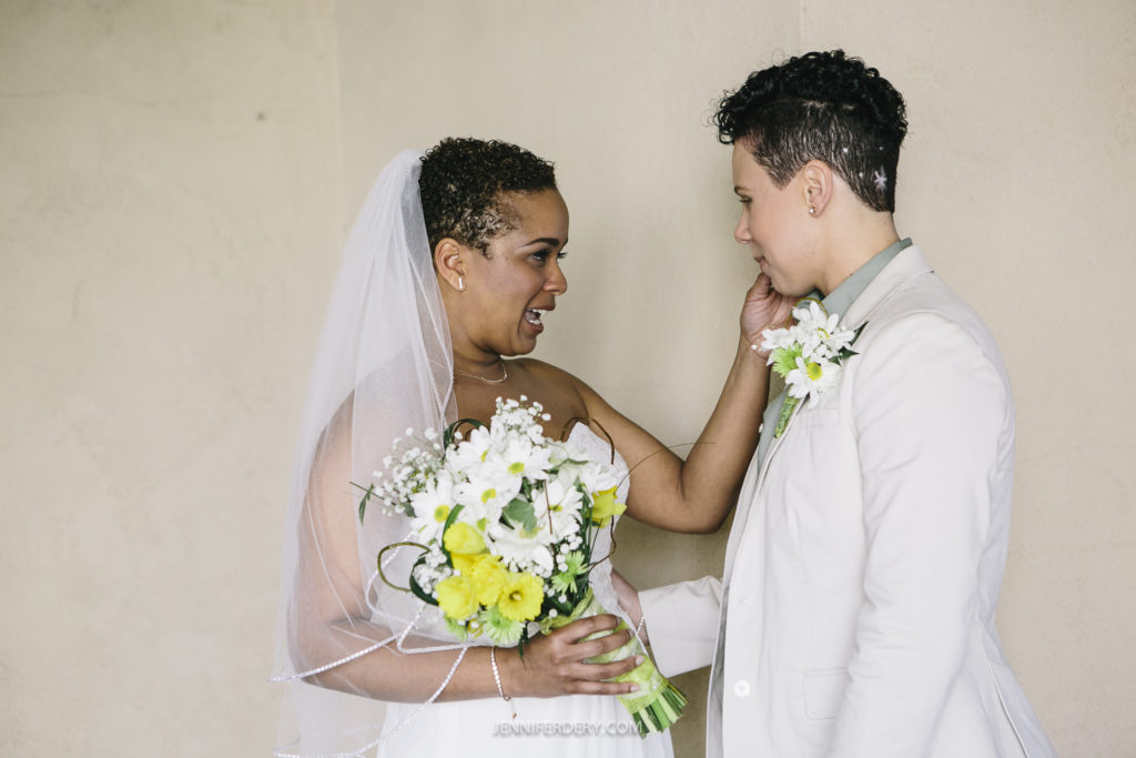 A couple, one in a wedding dress holding a bouquet of white and yellow flowers and the other in a light-colored suit, share an intimate moment at their Balboa Park wedding. They stand facing each other, smiling, as one gently touches the other's face.