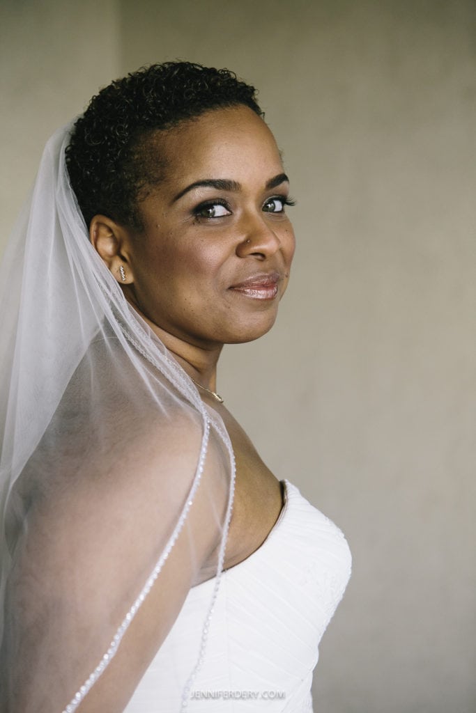 A person in a white wedding dress with a sheer veil over their short hair gazes at the camera. The neutral background focuses attention on their calm expression and smile, capturing the serene essence of a Balboa Park wedding.