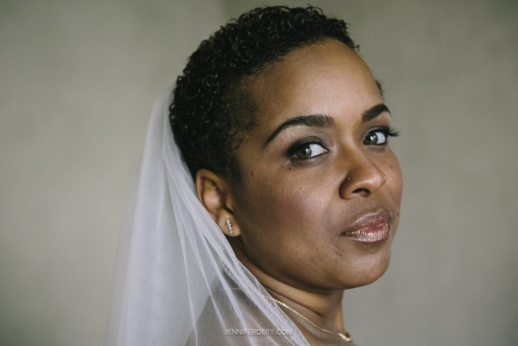A close-up of a bride with short curly hair, wearing a white veil. She is looking at the camera with a calm and composed expression, showcasing her elegant makeup and subtle jewelry. The background of Balboa Park is blurred, drawing focus to her face.