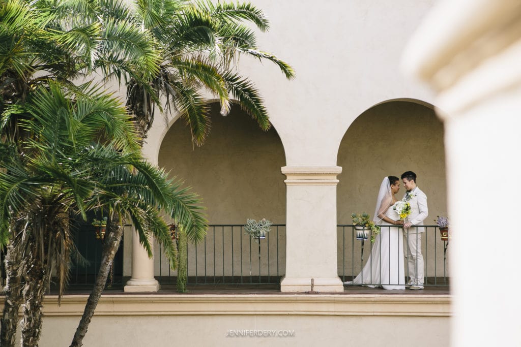 A newlywed couple stand on a balcony in Balboa Park, framed by elegant arches and a cream-colored wall. One person is in a white gown holding a bouquet, and the other is in a light-colored suit. Lush green palm trees are visible on the left side of the image.