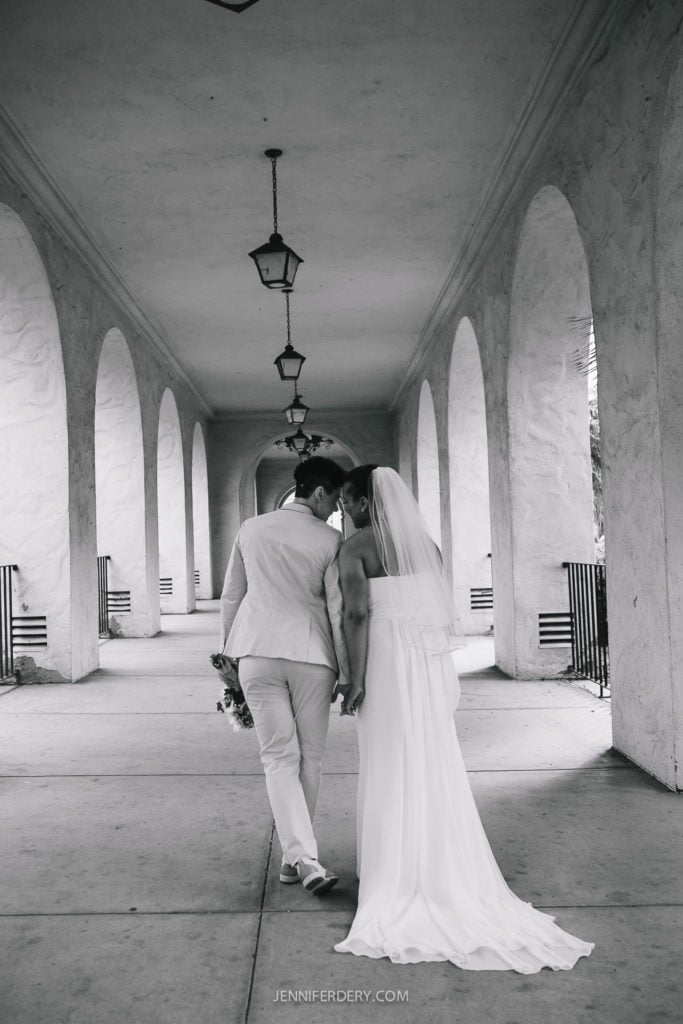 A newlywed couple walks hand in hand down a hallway with arches on either side at their Balboa Park wedding. One person wears a light-colored suit, and the other wears a flowing white gown with a veil. They share an intimate moment, touching heads and holding a bouquet in one hand. The setting appears elegant and timeless.