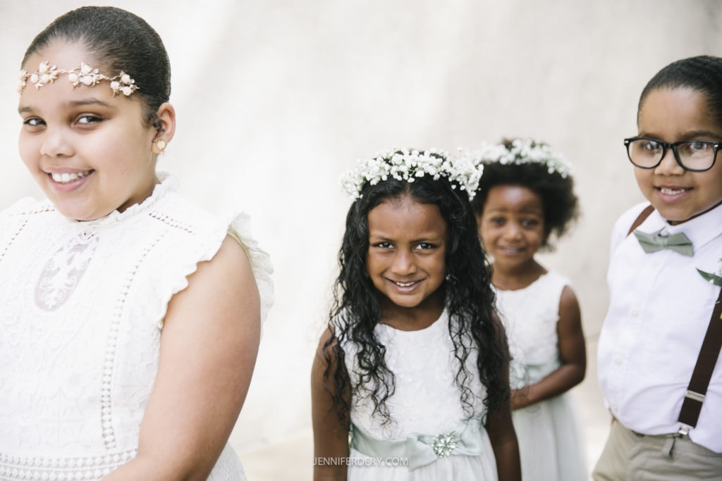 Four children dressed in formal attire pose for a picture at a Balboa Park wedding. Three girls wear white dresses, with two girls adorned in floral headpieces, while one boy wears glasses, a grey bow tie, and suspenders. They are standing in front of a light-colored background.