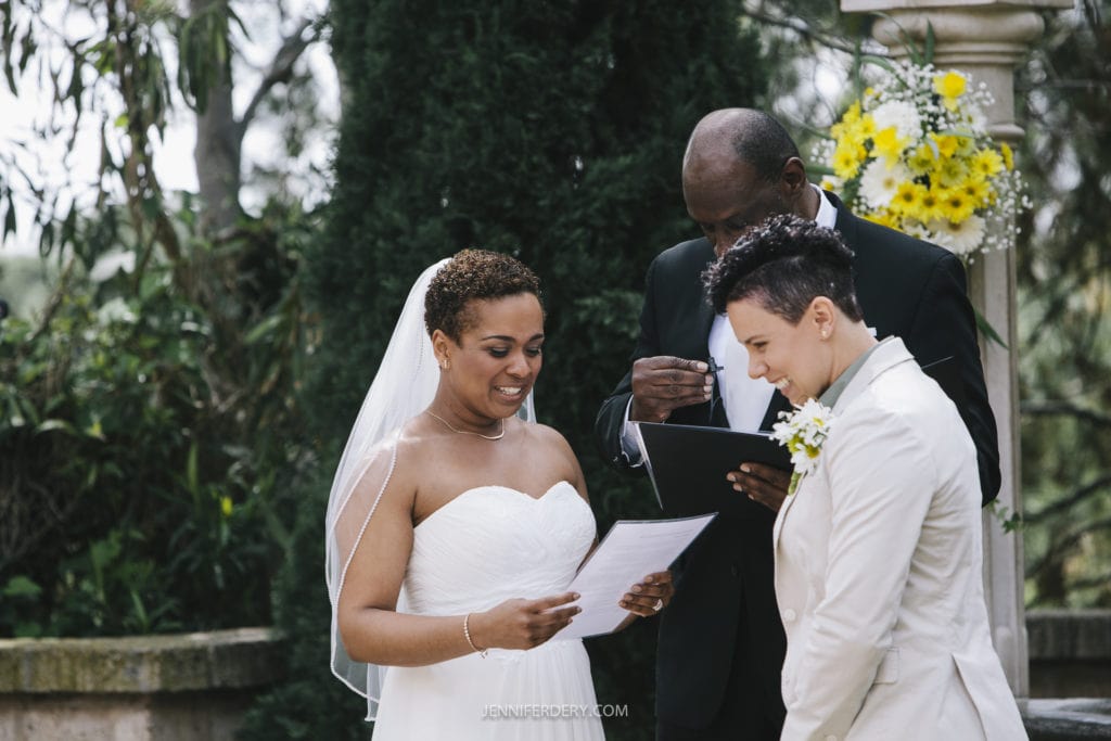 Two individuals are getting married outdoors in a beautiful Balboa Park wedding. Both are standing in front of an officiant who is reading from a book. One person is wearing a white dress and veil, holding a piece of paper and smiling. The other is in a light-colored suit, also smiling. Yellow flowers brighten the background.