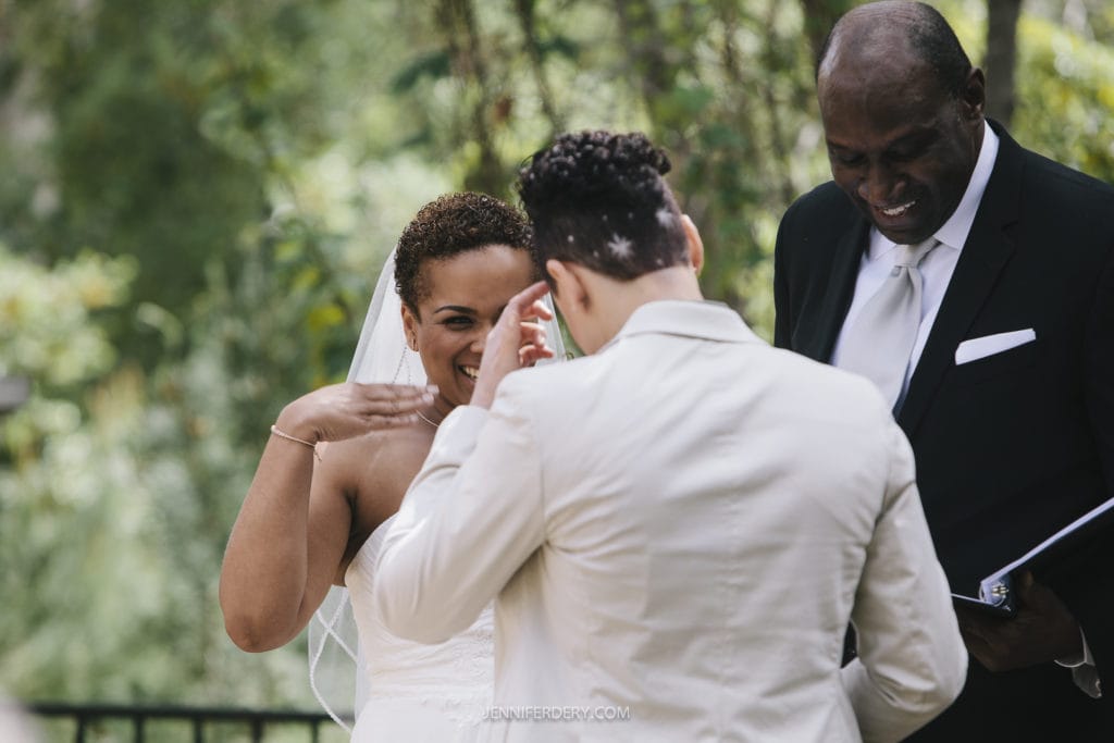 A newlywed couple stands together smiling, with the bride wiping tears of joy. The groom, in a white suit with a short haircut, and the bride, in a strapless wedding dress with a veil, are radiant at their Balboa Park wedding. An officiant in a dark suit and white tie is laughing beside them.