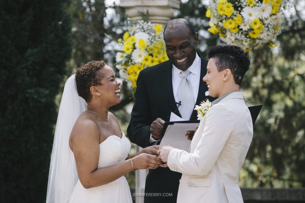 A joyous moment at an outdoor Balboa Park wedding. A smiling bride in a white dress and veil holds hands with her partner in a white suit, while an officiant in a black suit and white tie stands behind them, holding a book. Bright flowers decorate the background.