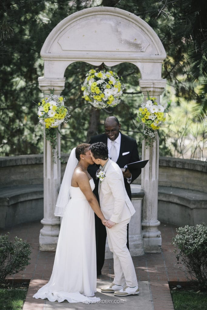 A couple shares a kiss at their wedding ceremony in Balboa Park, standing under a decorated stone arch adorned with floral arrangements. The person officiating the ceremony, holding a book, smiles in the background. The surroundings are lush with greenery.