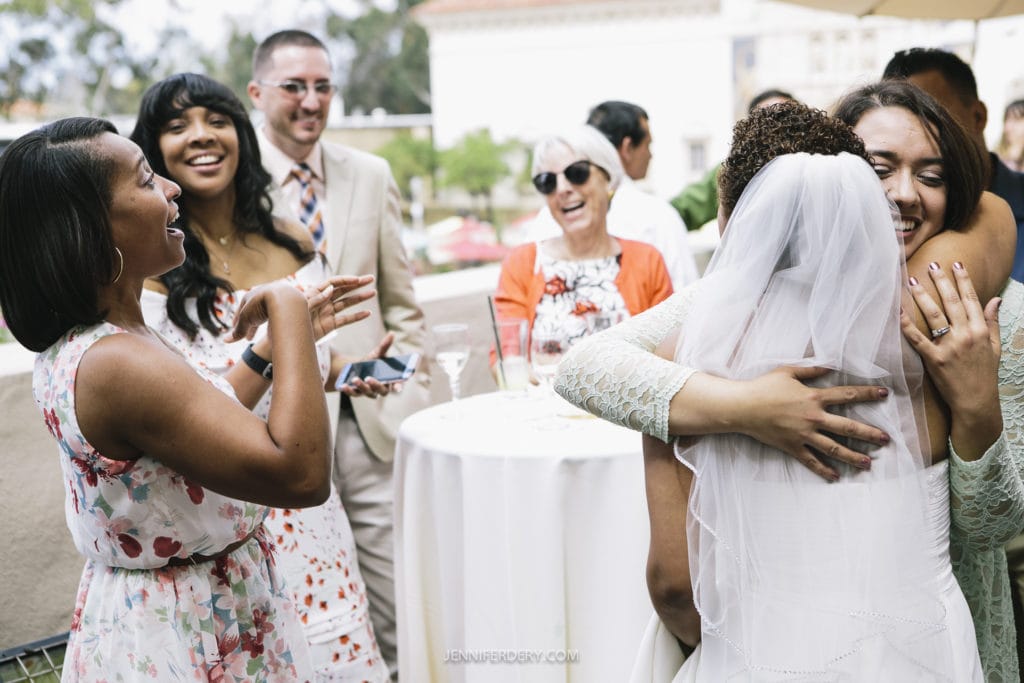 A group of people at a Balboa Park wedding reception, with a bride in a veil hugging a guest. Everyone is smiling and appearing joyful. Some are wearing summer dresses, and one person is in a suit. A round table and another guest wearing sunglasses are visible in the background.