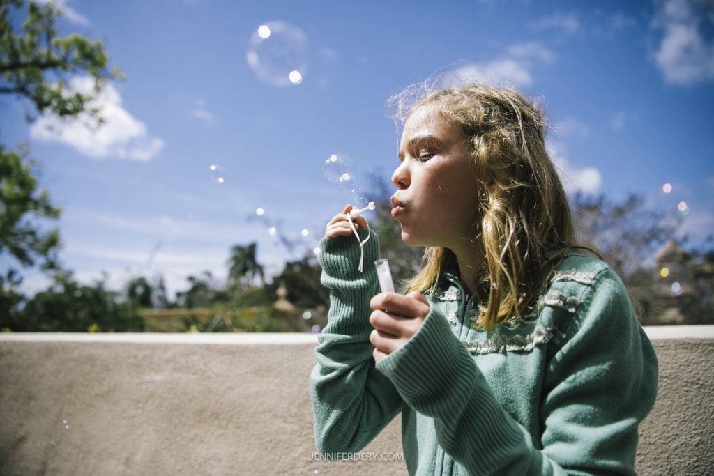 A young girl with blonde hair wearing a green sweater blows soap bubbles against a clear blue sky at Balboa Park. Trees and buildings are blurred in the background, highlighting the bubbles, capturing the magic of a perfect moment akin to a wedding day.